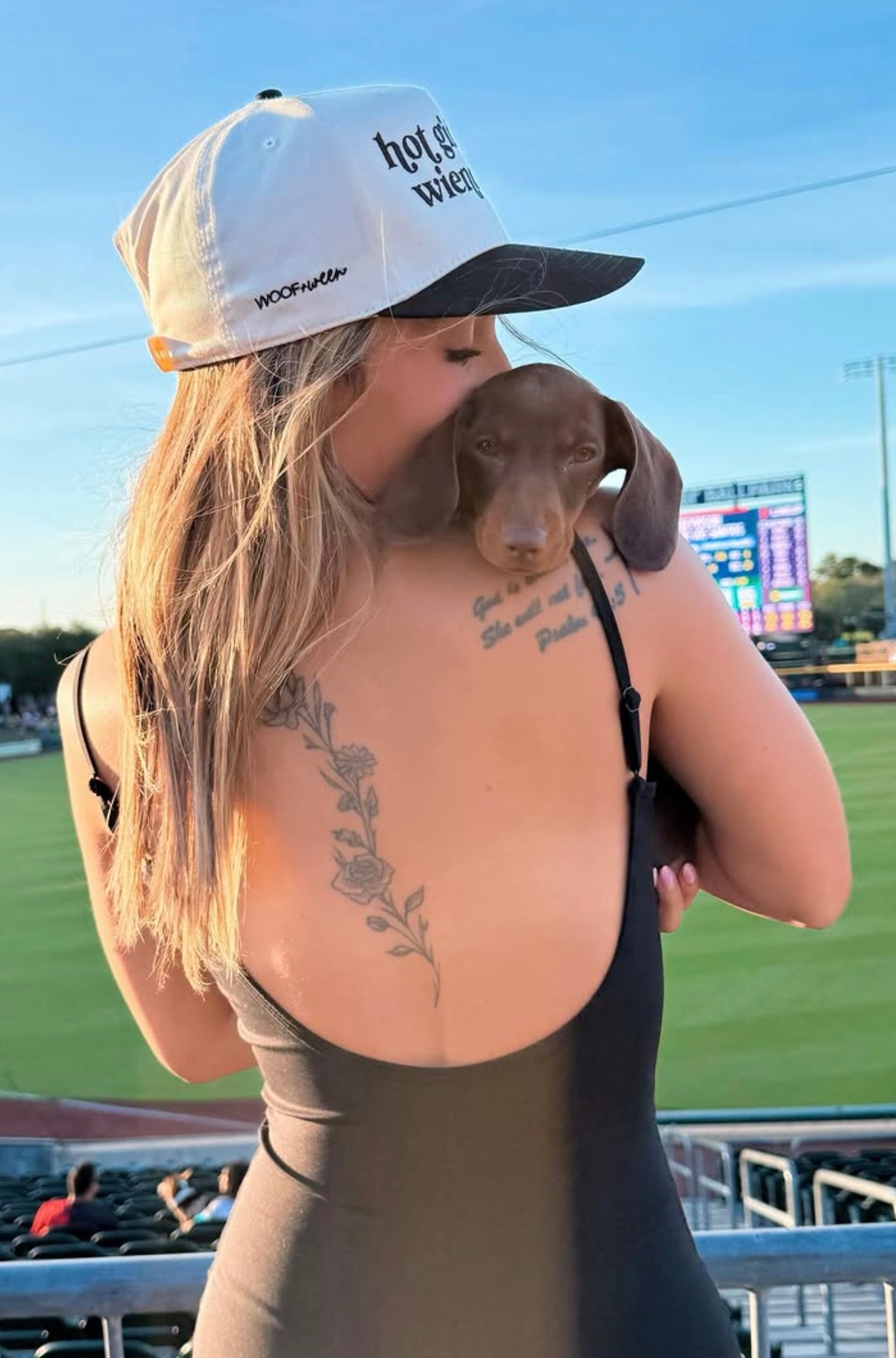 A woman at a baseball game wearing a white and black woof+ween trucker hat with 'hot girls have wiener dogs” embroidered on it