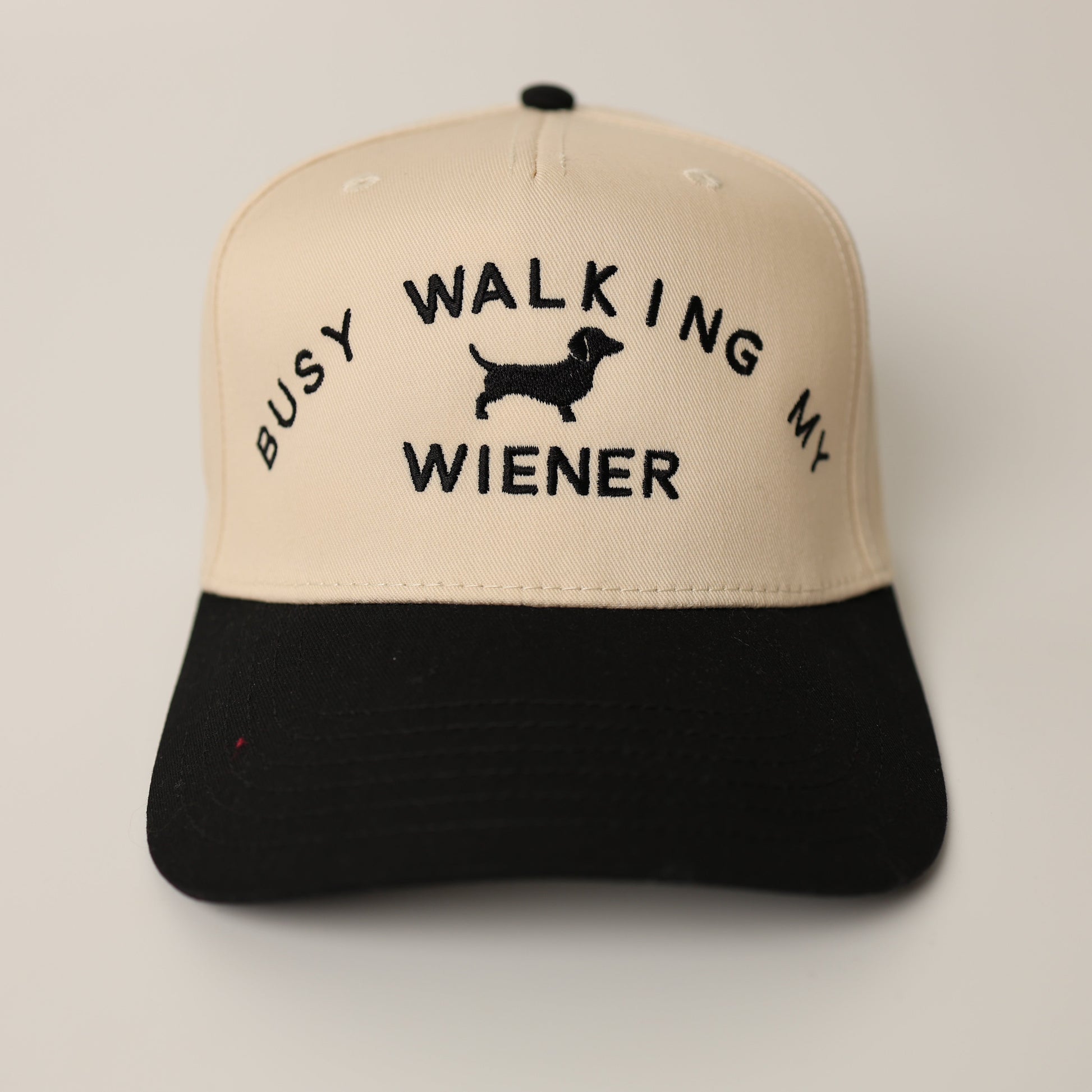 Beige and black cap with 'Busy Walking My Wiener' text on a light gray background