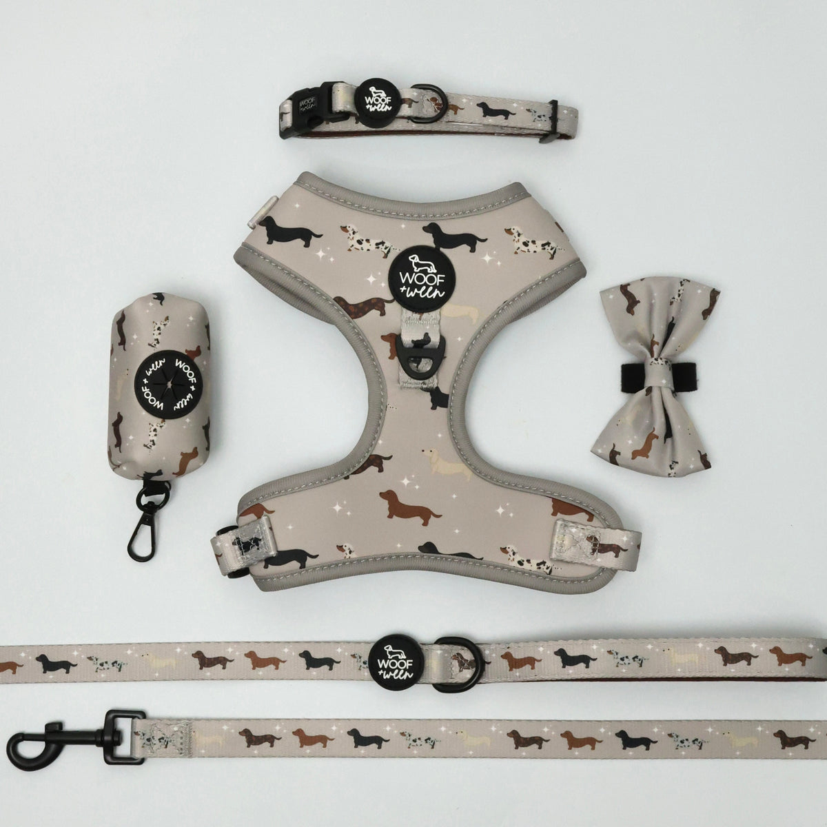 A dog harness set laid out on a white background, including a harness, leash, collar, and matching fabric accessories with a beige base and black and brown dachshund design illustrations.