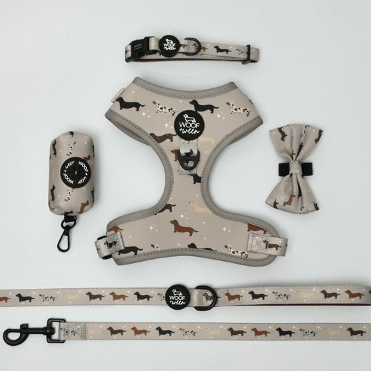 A dog harness set laid out on a white background, including a harness, leash, collar, and matching fabric accessories with a beige base and black and brown dachshund design illustrations.