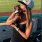 A woman at a baseball stadium wearing a woof+ween black and tan baseball cap with the text 'hot girls have wiener dogs' while holding her dachshund