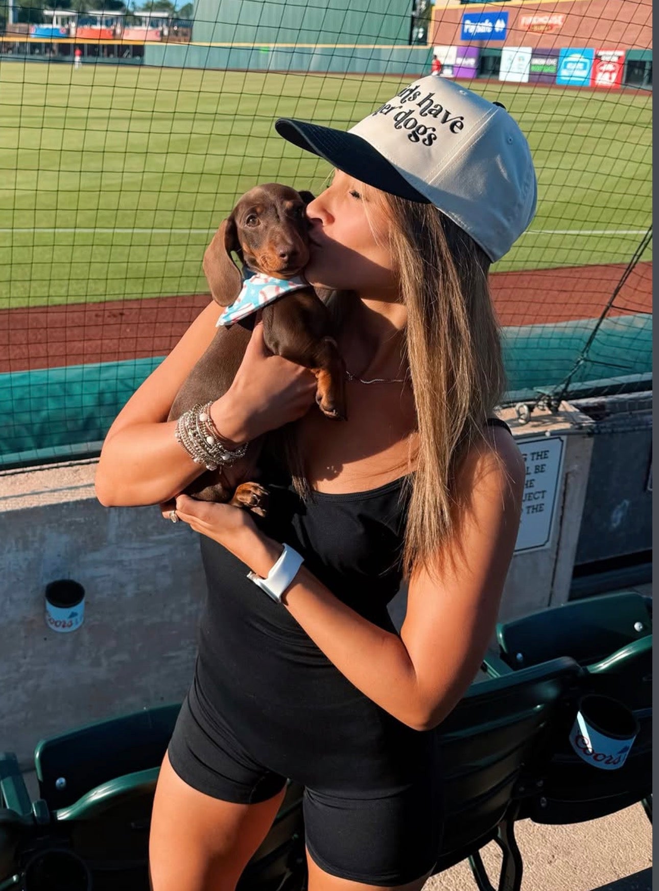 A woman at a baseball stadium wearing a woof+ween black and tan baseball cap with the text 'hot girls have wiener dogs' while holding her dachshund